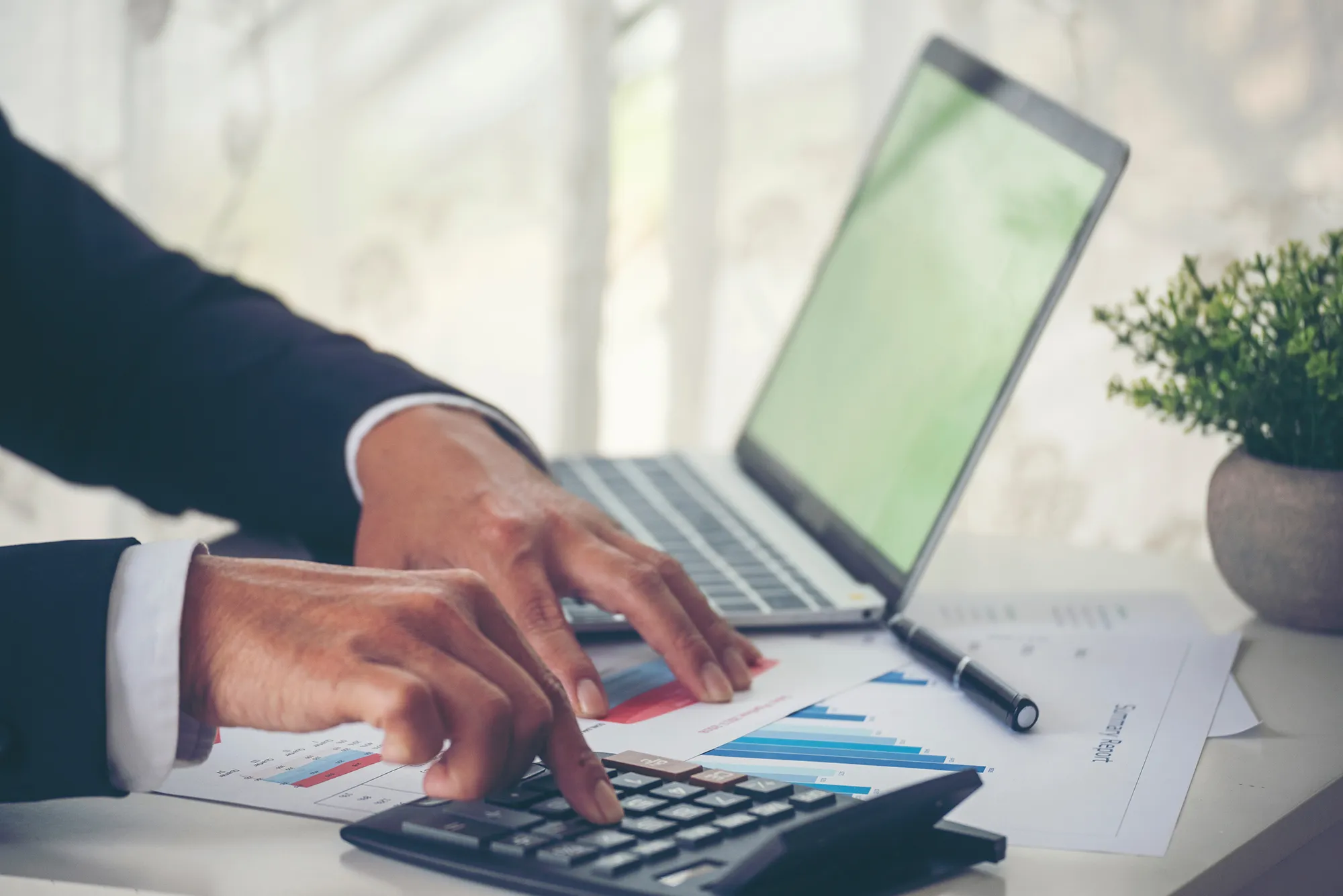 Hands of a man with a laptop and calculator on a desk, pen, papers and plant pot in background