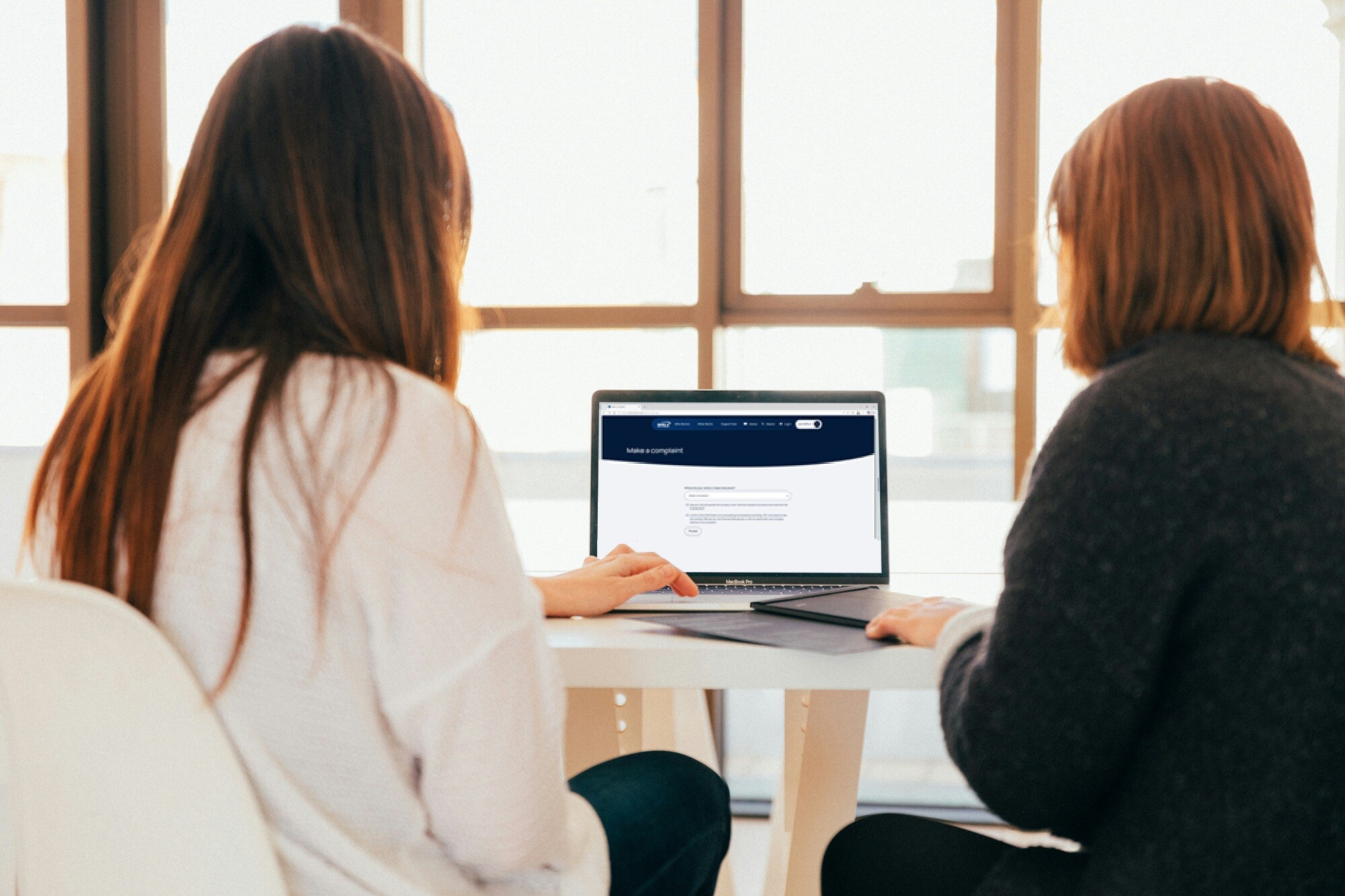 Two women sit at a white table looking at one laptop. They are working collaboratively to browse the B V R L A site and use the A D R service