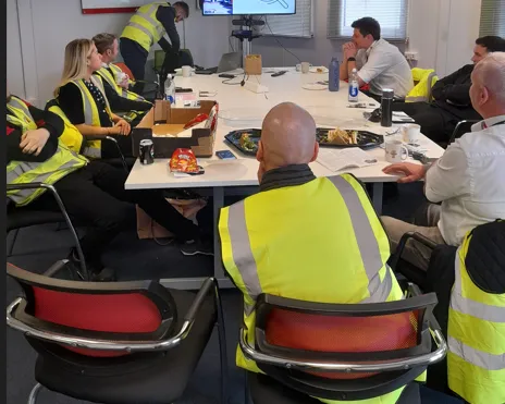 A group of people, most wearing high visibility jackets are sitting around a table in a training room