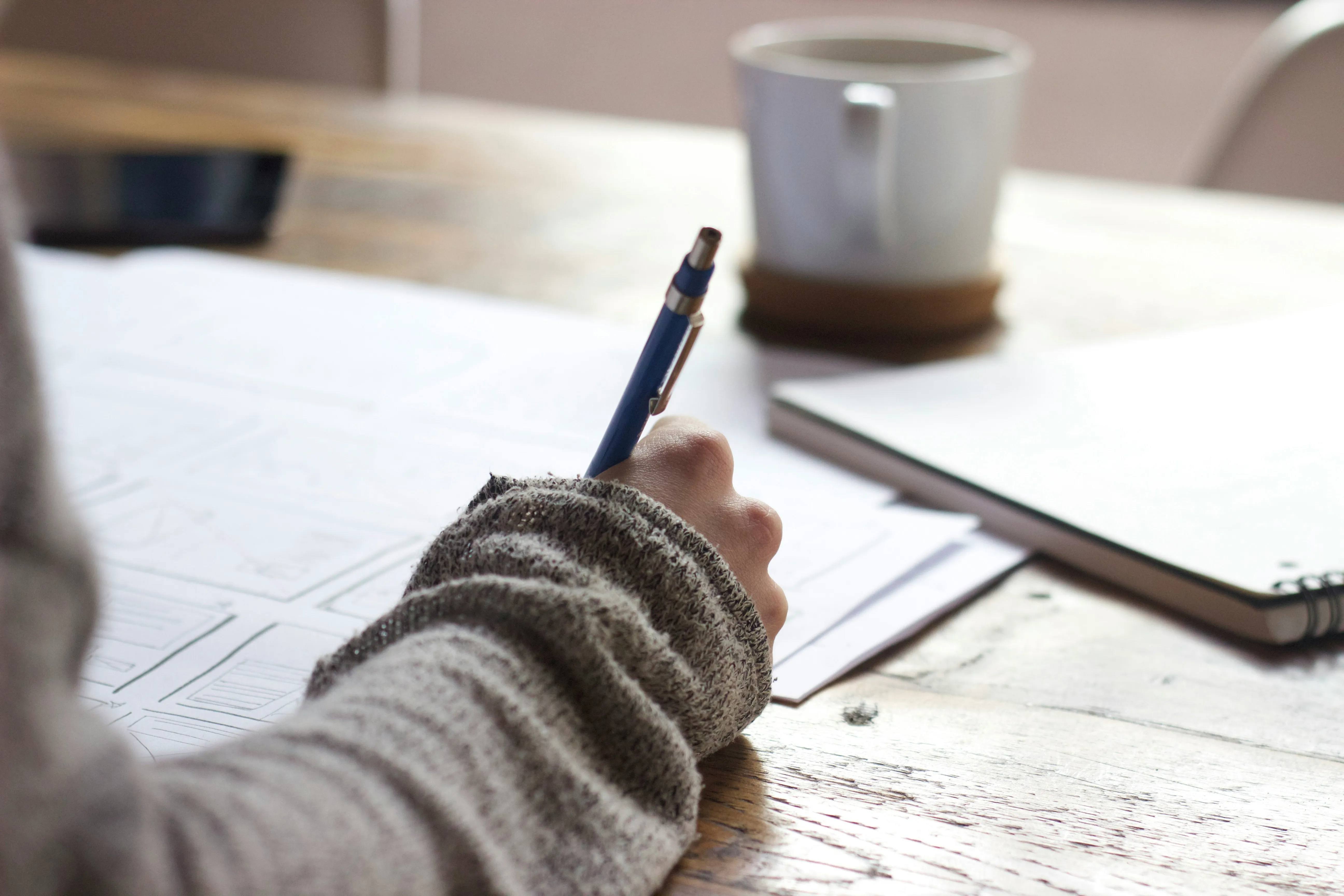 A person writing on paper at a table with a white mug