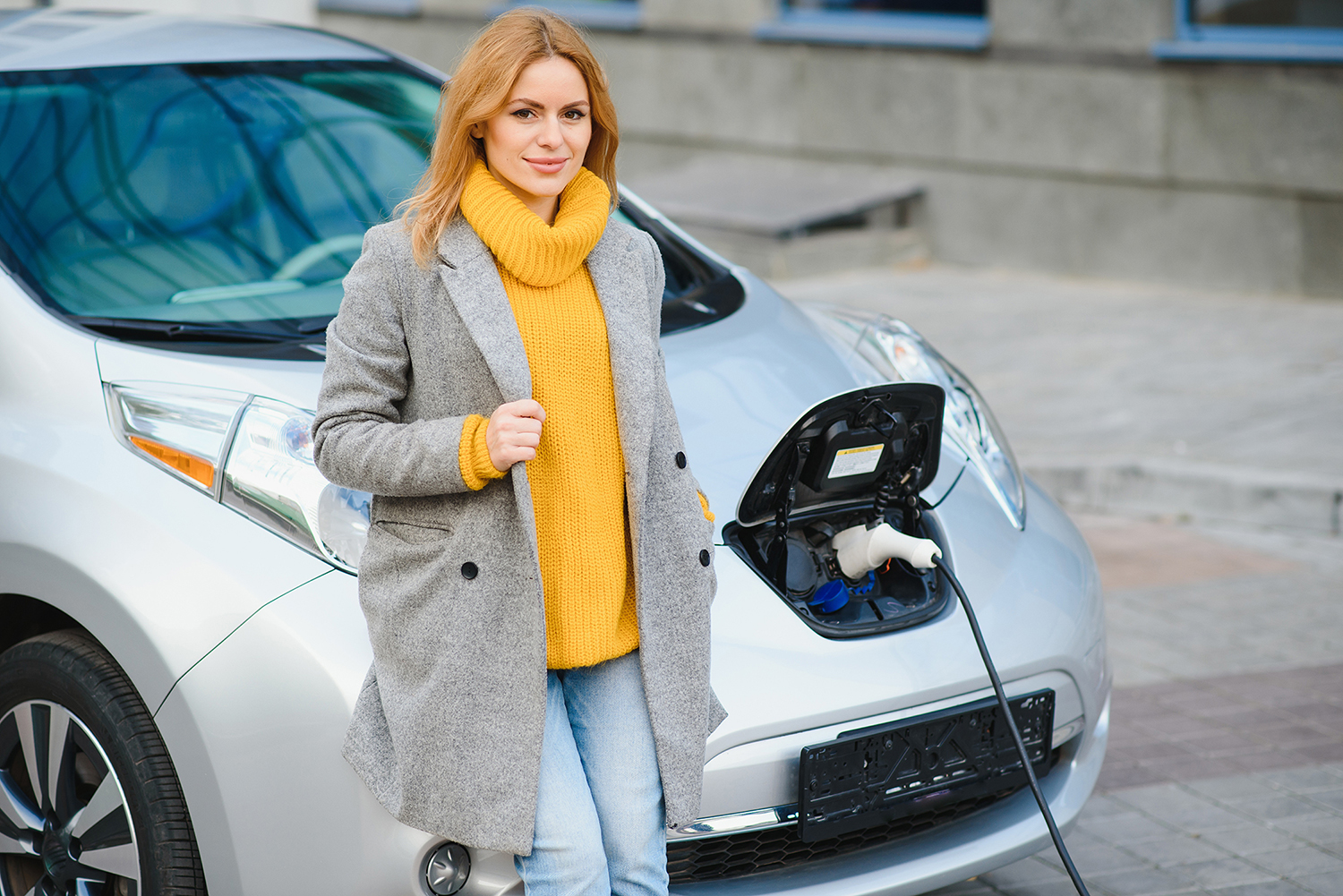 Lady in yellow jumper and grey coat leaning on a silver electric vehicle whilst it is charging