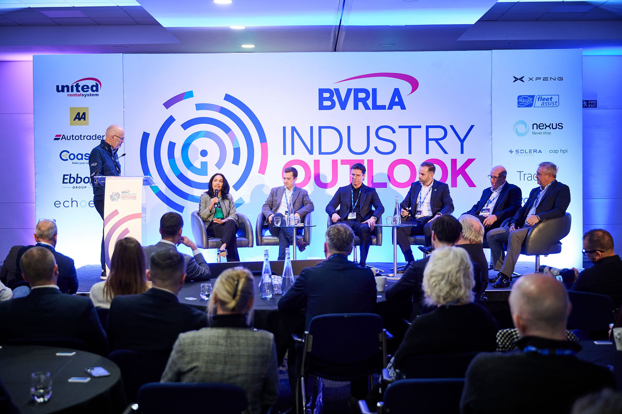 The stage at the Industry Outlook Conference with the panel of senior leaders and Toby Poston standing to left as chair. Industry Outlook branding in background