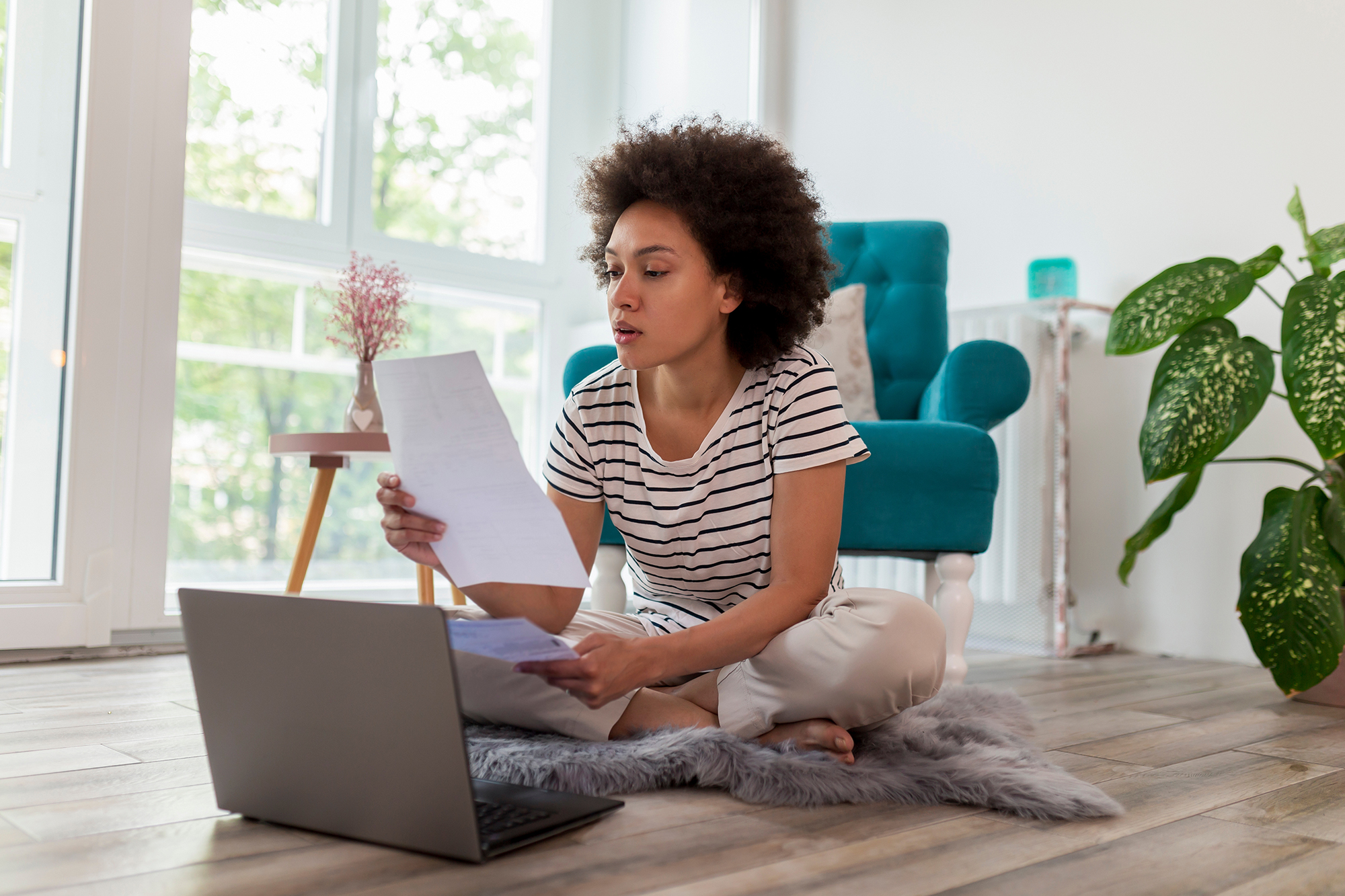 Woman sitting on floor with laptop and holding papers, looking concerned, room in background with blue chair and large plant