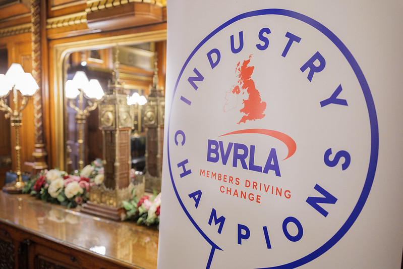 Image shows the Industry Champions logo - a blue and red speech bubble with a map of the UK inside, in front of a gold model of Big Ben, taken inside the Houses of Parliament